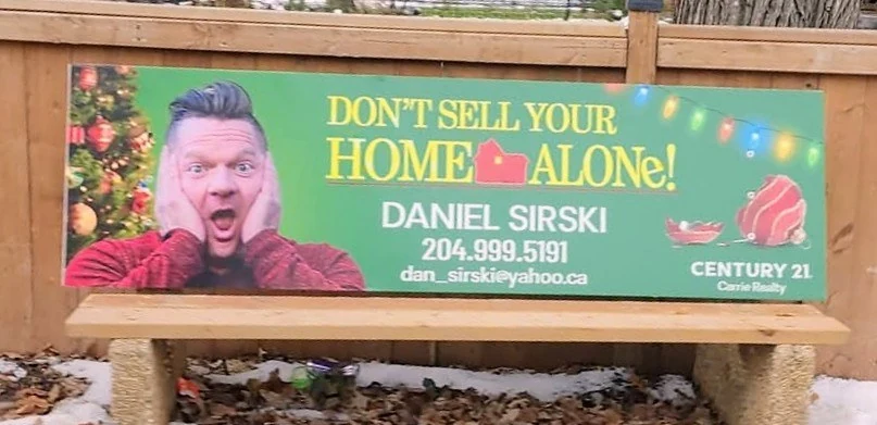 A green holiday-themed bus bench advertisement with Daniel Sirski holding his hands to his cheeks beside the line “Don’t sell your home alone”, framed by a decorated tree and string lights