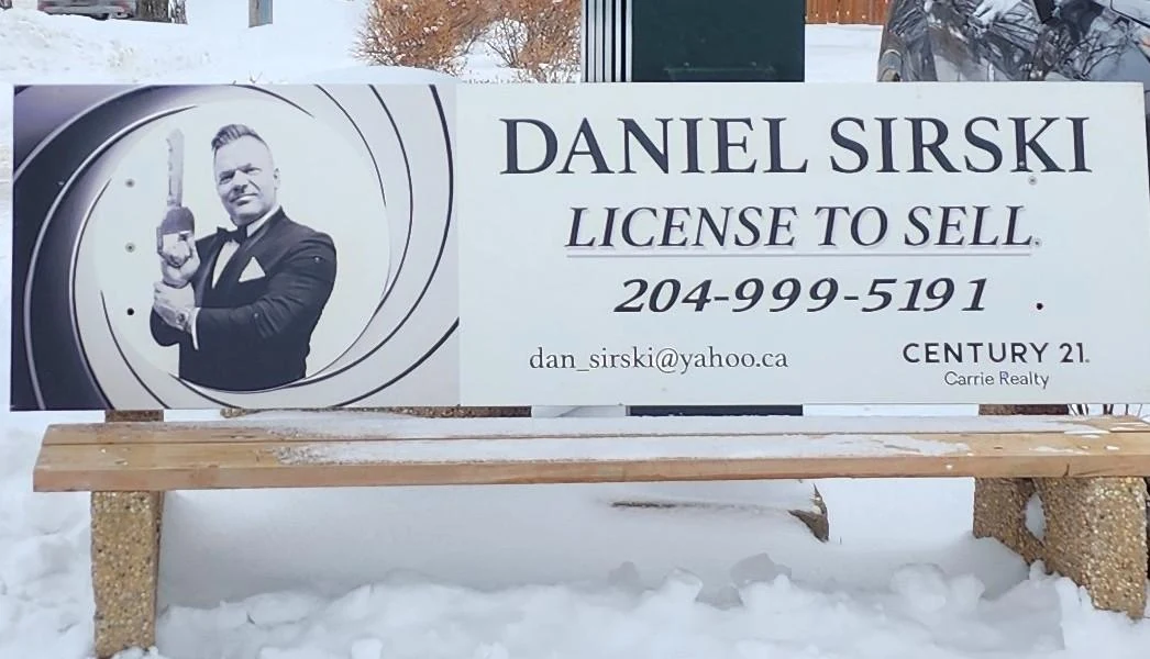 A Winnipeg bus bench advertisement in a snowy setting showing Daniel Sirski in a tuxedo holding a house key inside a gun-barrel graphic, beside his name and phone number