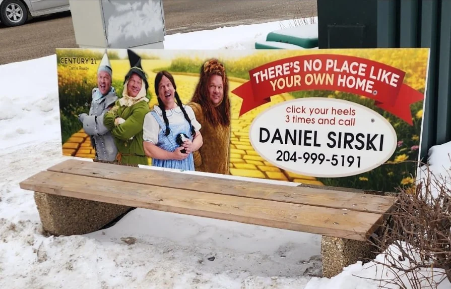 A bus bench advertisement on a snowy Winnipeg street showing four costumed characters on a yellow brick road beside the line “There’s no place like your own home” and Daniel Sirski’s name